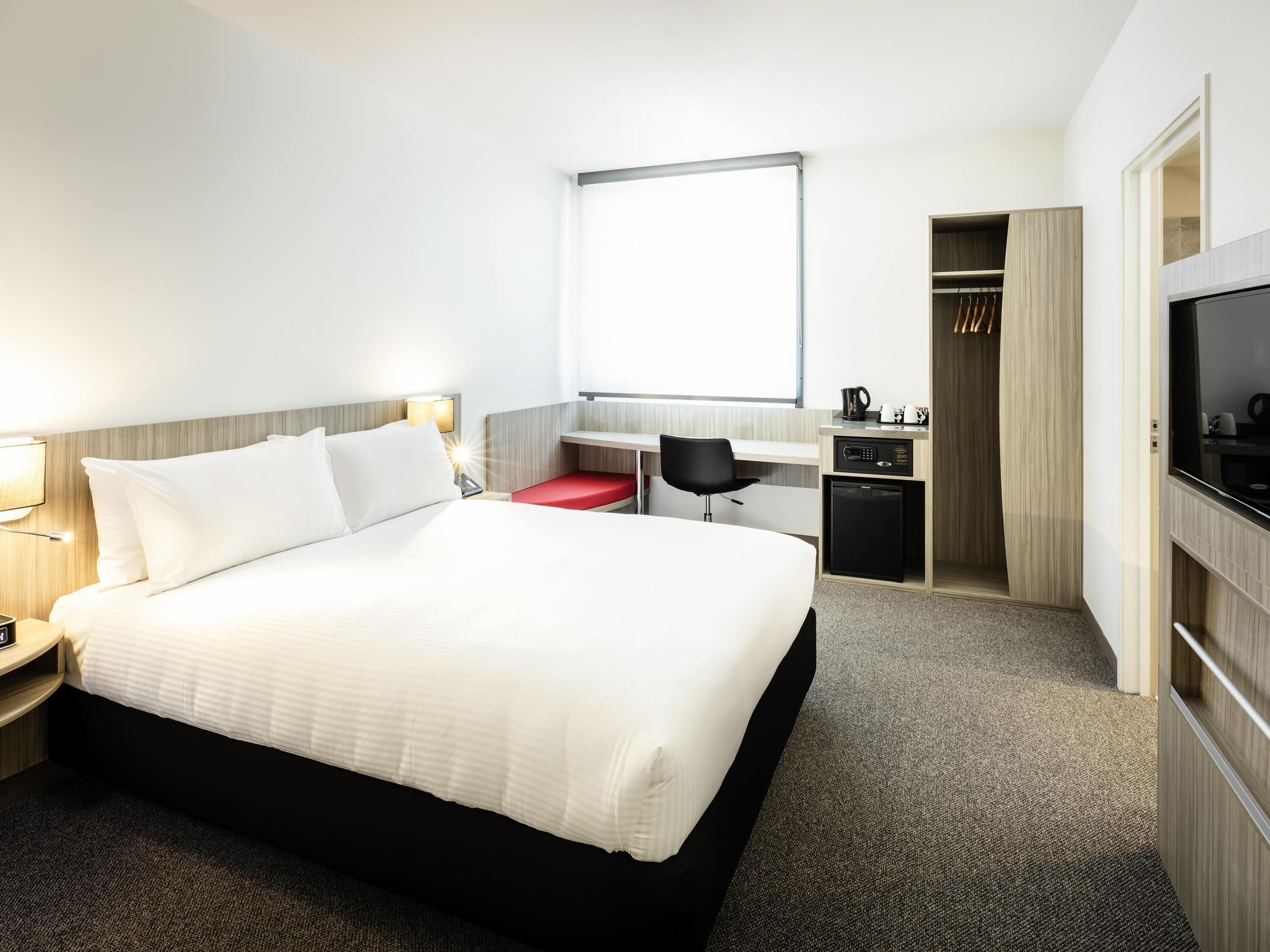 A hotel room with a bed, desk, chair, TV, wardrobe, and a small kitchenette.