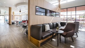 A modern lounge area with a sofa, armchairs, and a coffee table. There is a large digital display on the wall and a view of the outside through large windows. - ibis Mackay (South Mackay)