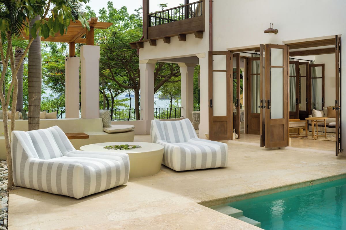 Un área junto a la piscina con dos sillones rayados, una mesa redonda y una pérgola de madera.