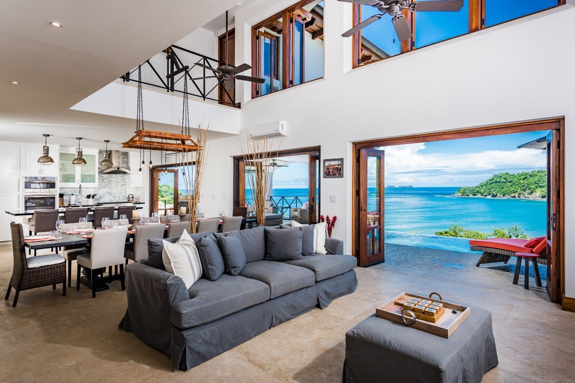 A spacious living area with a gray sofa, dining table, and chairs. A kitchen is visible in the background, and there are large windows offering a view of the ocean.