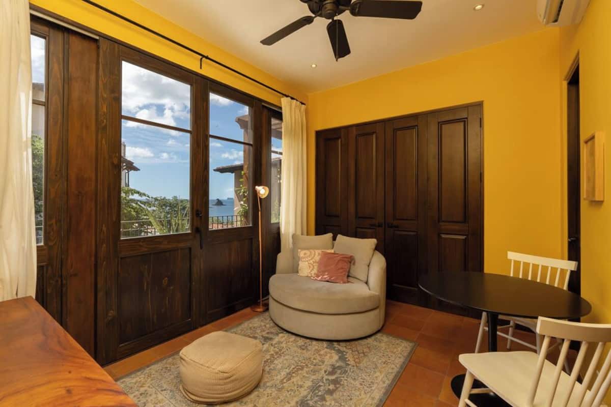 Una habitación con un ventanal grande, un ventilador de techo, un sofá redondo, una mesita y una silla.