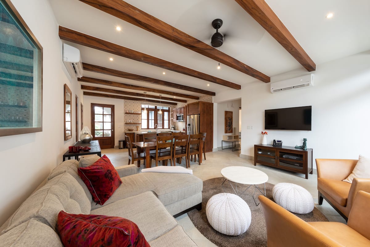 Amplia sala de estar con sofá, otomanas, televisor y zona de comedor con mobiliario de madera.