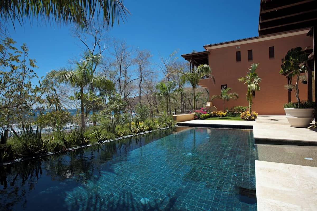 Una piscina azul cristalina con borde de azulejos, rodeada de plantas tropicales y un edificio de techo plano.