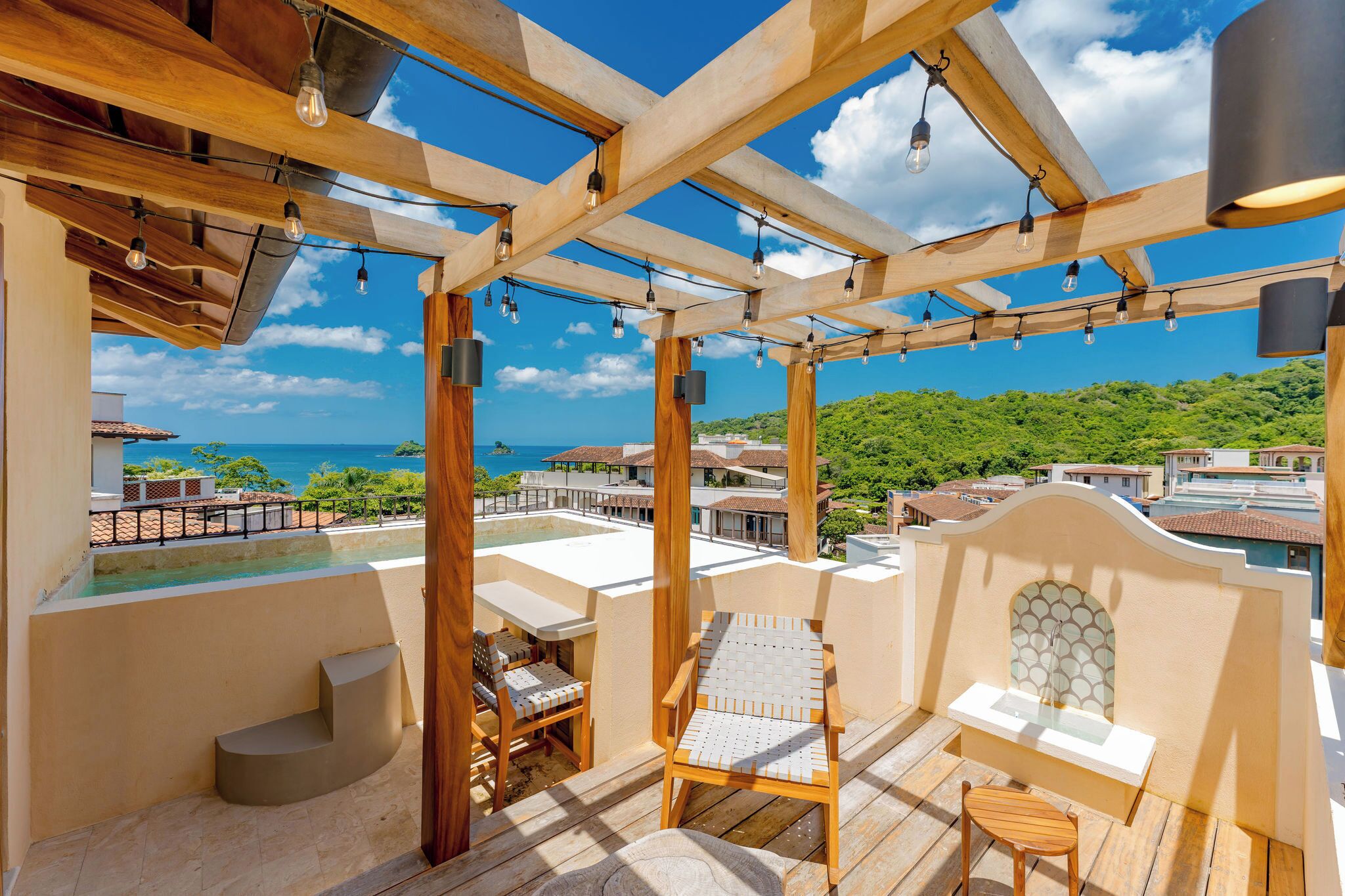 A wooden deck with a pool, outdoor seating, and a view of the ocean and greenery.