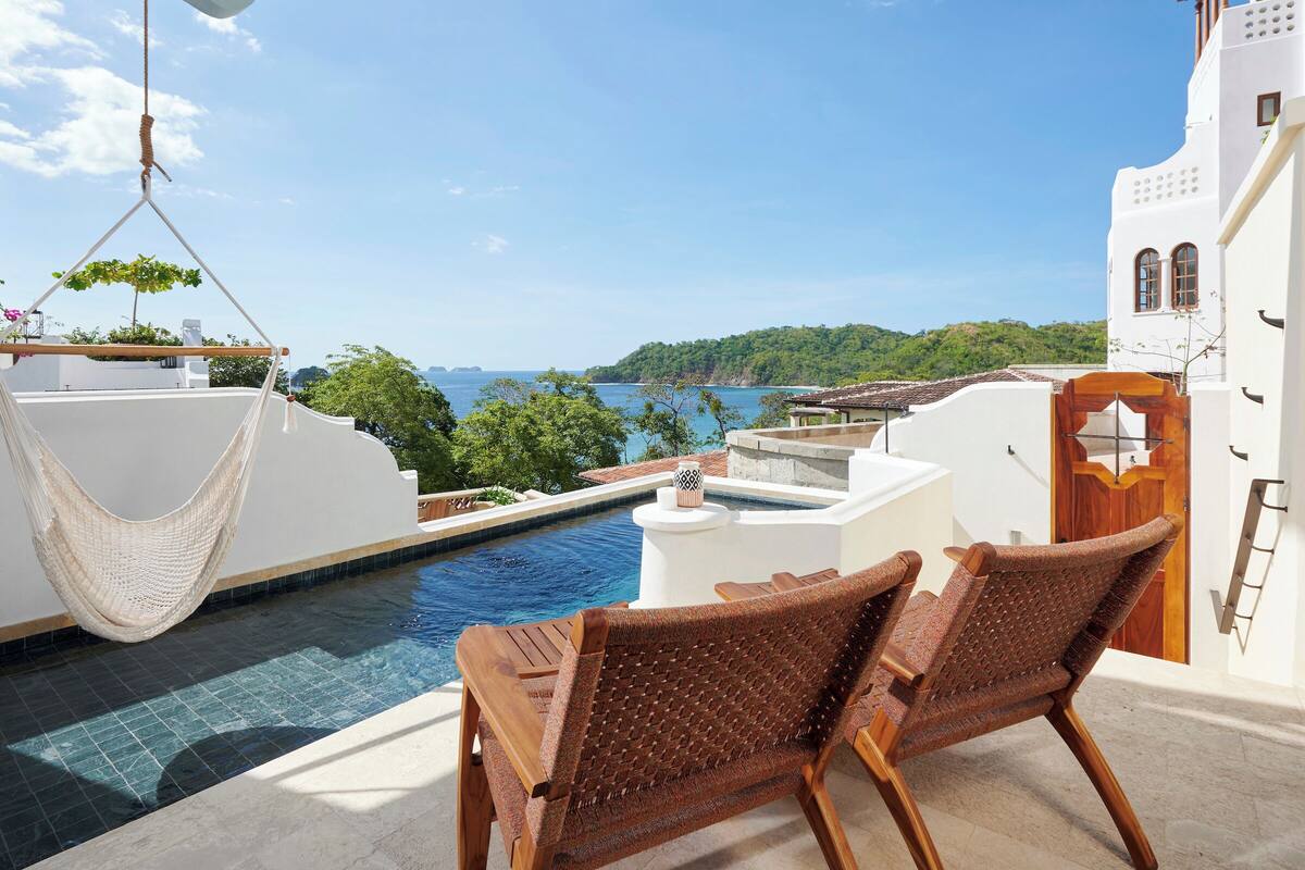 Zona junto a la piscina con una hamaca, dos sillas de mimbre y vista al mar.