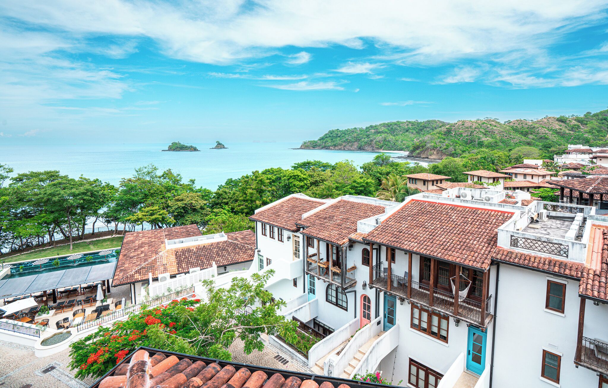 A coastal town with terracotta-roofed buildings, a pool area, and a view of the sea and islands.