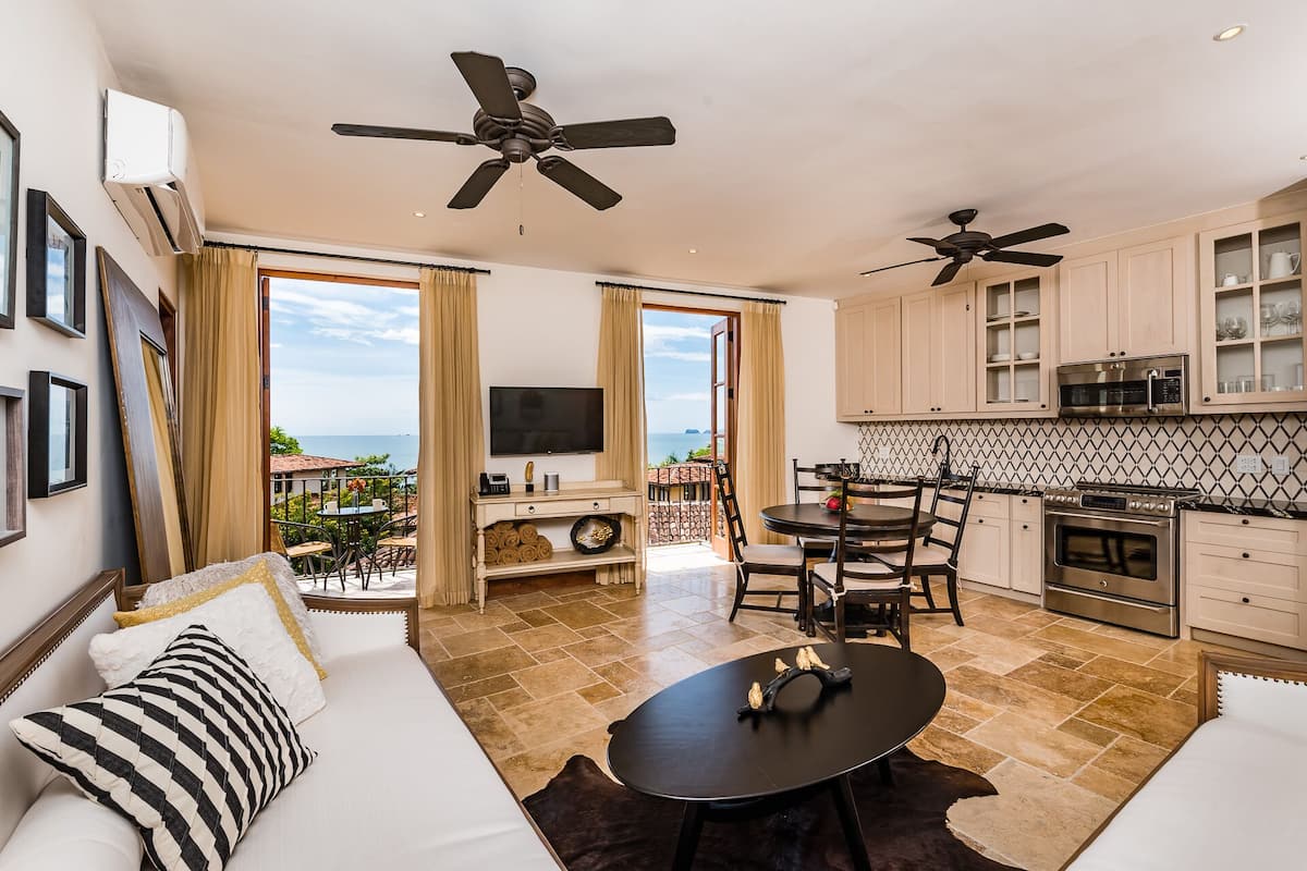 Una sala de estar moderna con un sofá, una mesa de centro y un televisor. Hay un balcón con vistas al mar. La cocina tiene armarios y un microondas.