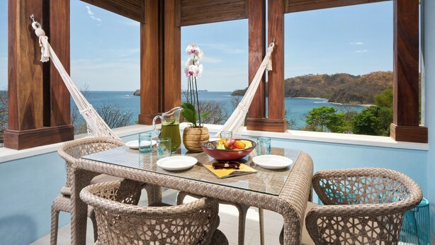 A glass-top table with wicker chairs, a fruit bowl, a pitcher, and a vase with flowers, overlooking a coastal view. - Las Catalinas Doorway (Tempate)