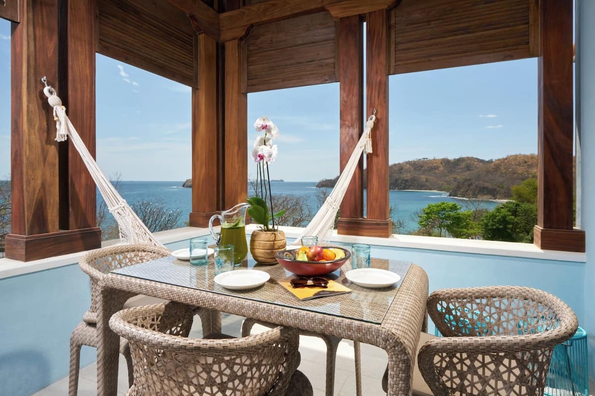 Mesa de vidrio con sillas de mimbre, frutero, jarra y un florero con flores, con vista al mar.