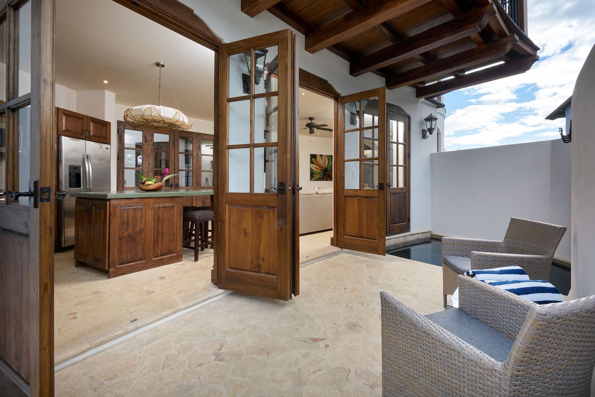 Una cocina amplia con armarios de madera y una isla grande, un comedor con mobiliario de mimbre y vista a una piscina.