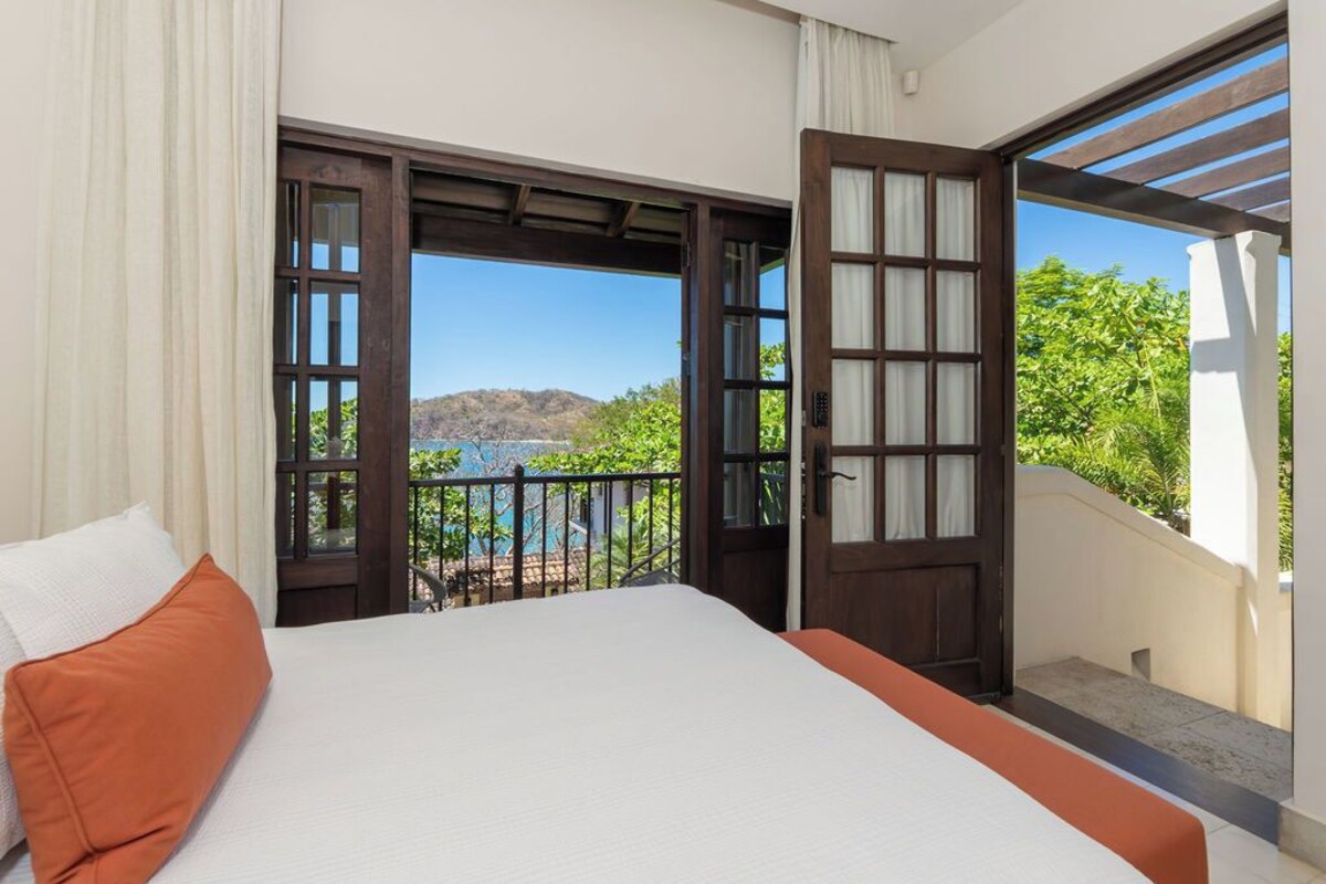 A bedroom with a large bed, an open door leading to a balcony with a view of the sea and hills.