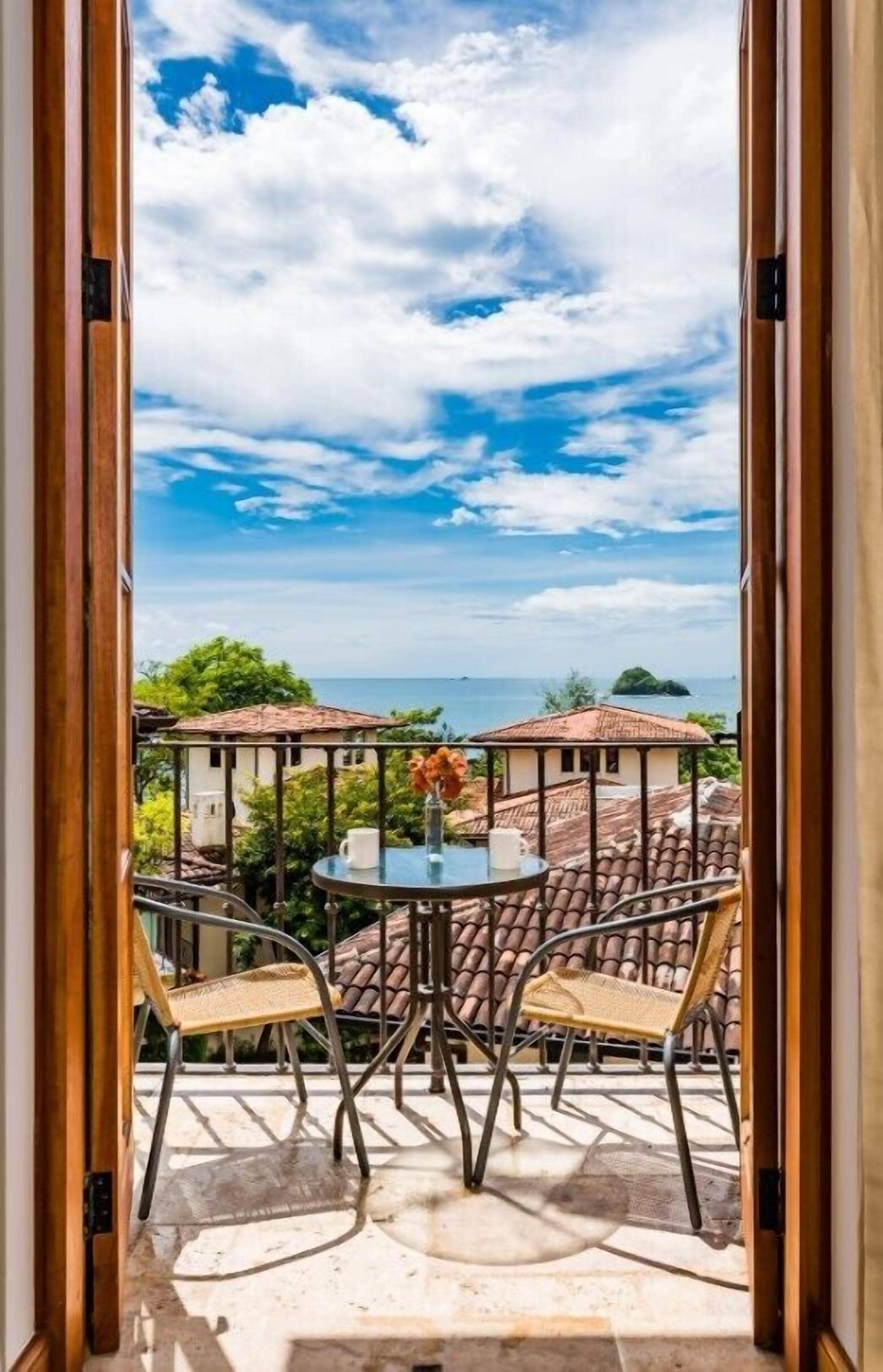 A balcony with a table and chairs, overlooking a coastal view with buildings and the sea.