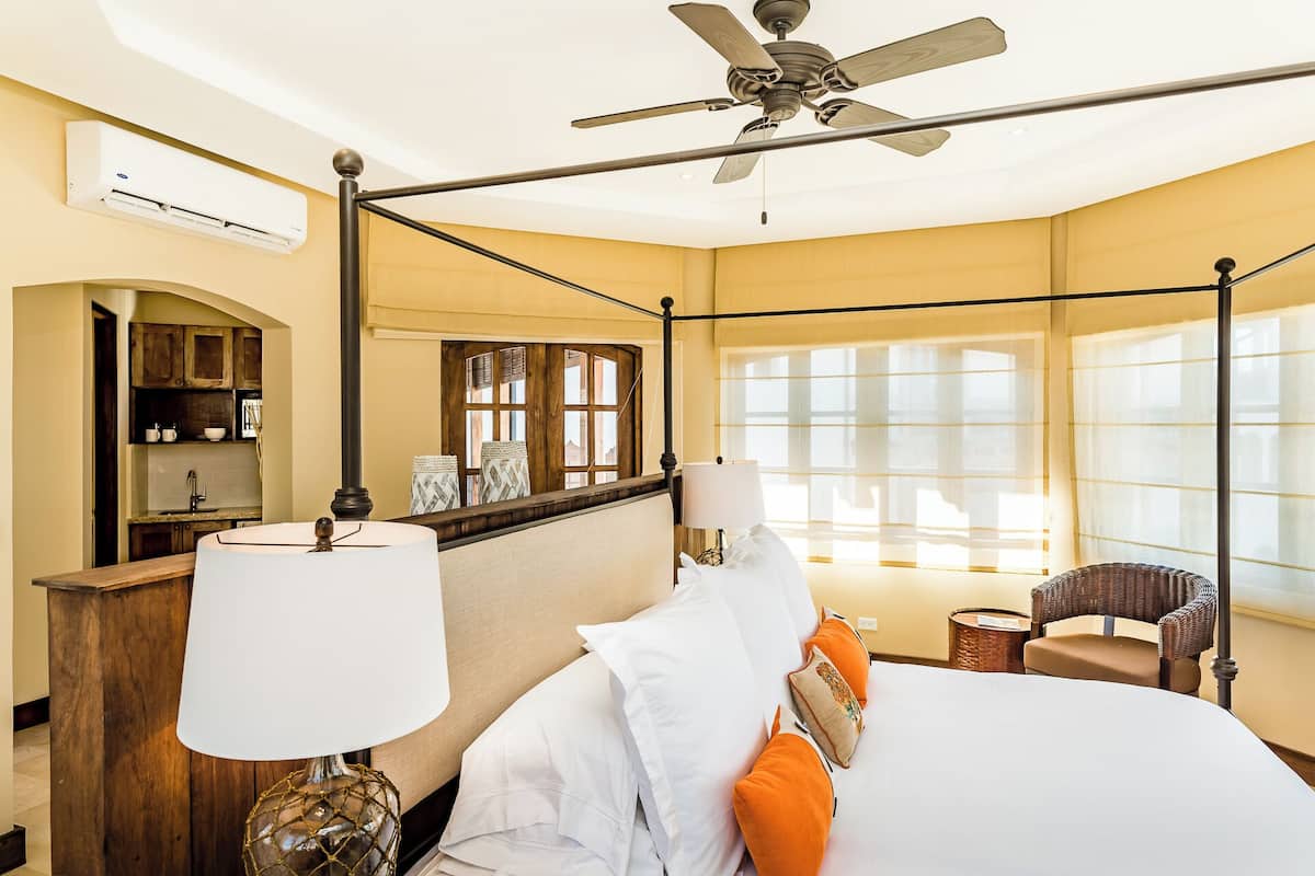 Habitación de hotel con cama con dosel, ventilador de techo, ventana grande con cortinas traslúcidas, una silla de mimbre y vista a la ciudad.