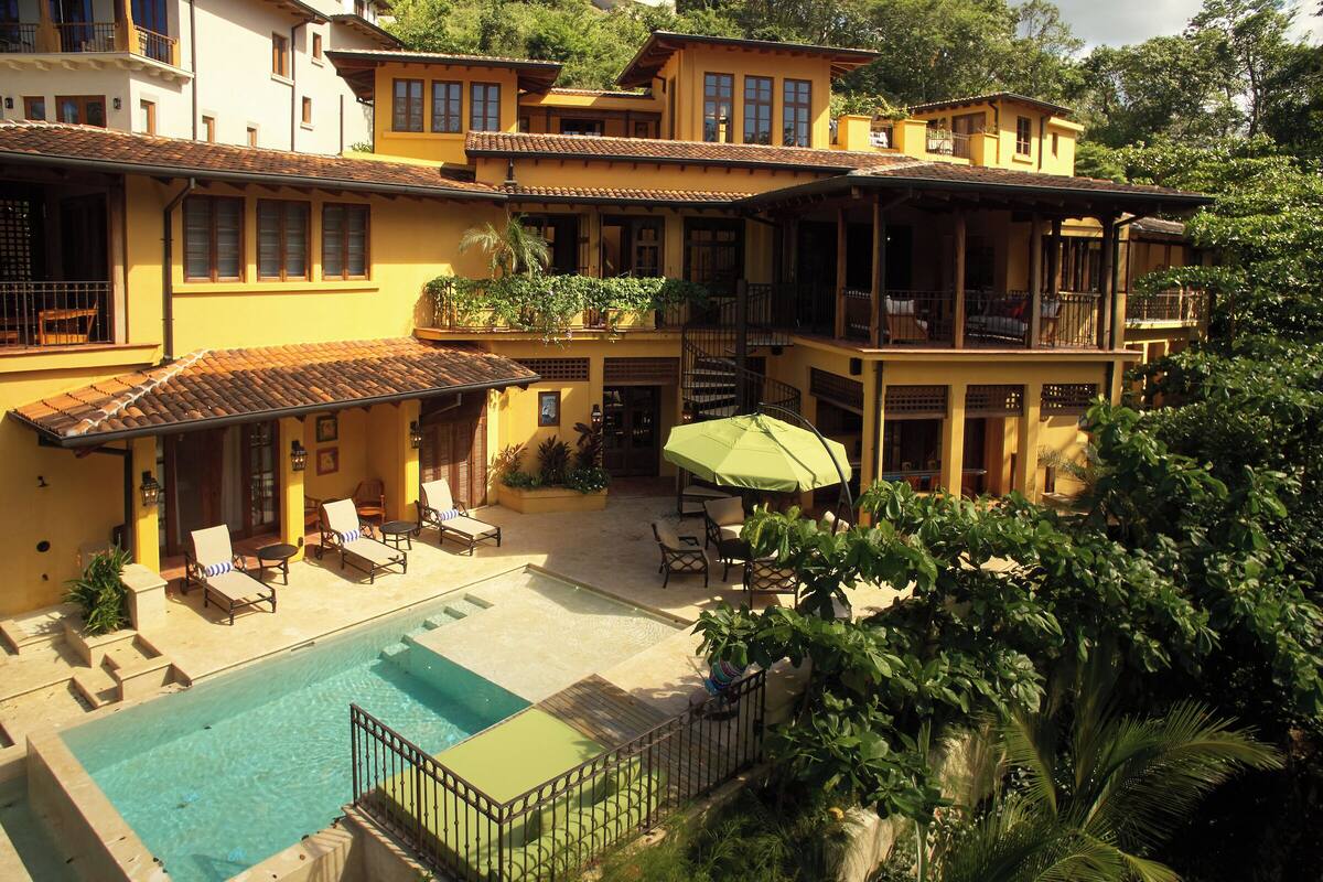 Un edificio amarillo con piscina, mobiliario exterior y un patio cubierto.