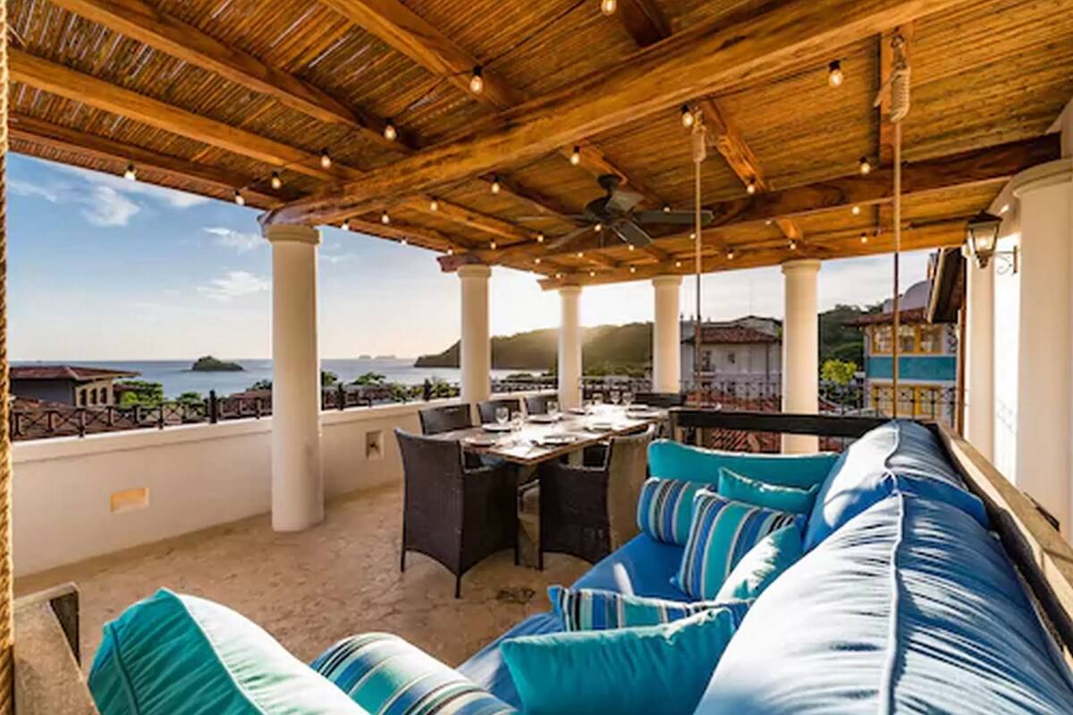 Un área de estar al aire libre con techo de madera, columnas blancas y vista al mar.