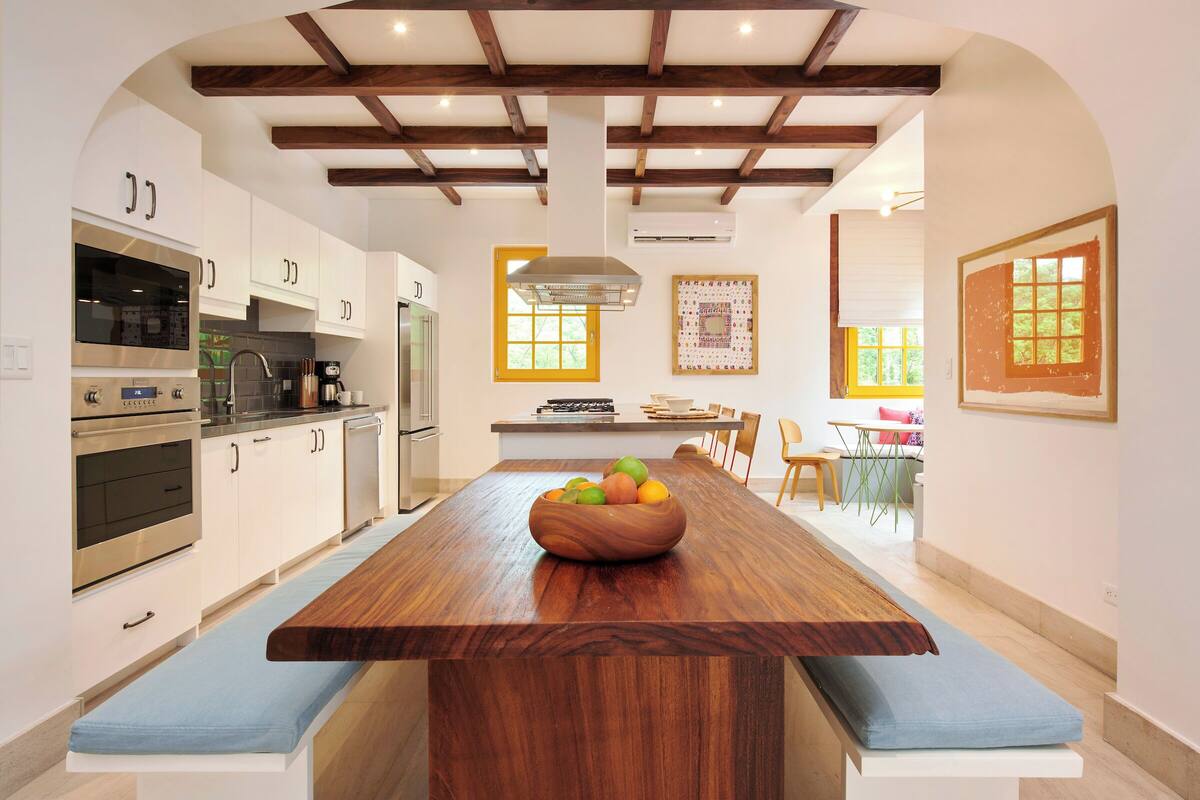 Una cocina moderna con armarios blancos, una isla de madera con un frutero, y un comedor con mesa y sillas.