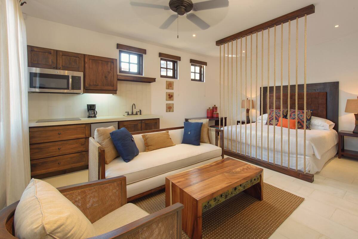 Una habitación de hotel moderna con kitchenette, una cama con cabecera de madera, un sofá, una mesa de centro y un ventilador de techo.