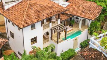 A two-story building with a tiled roof, a swimming pool, and surrounding greenery.