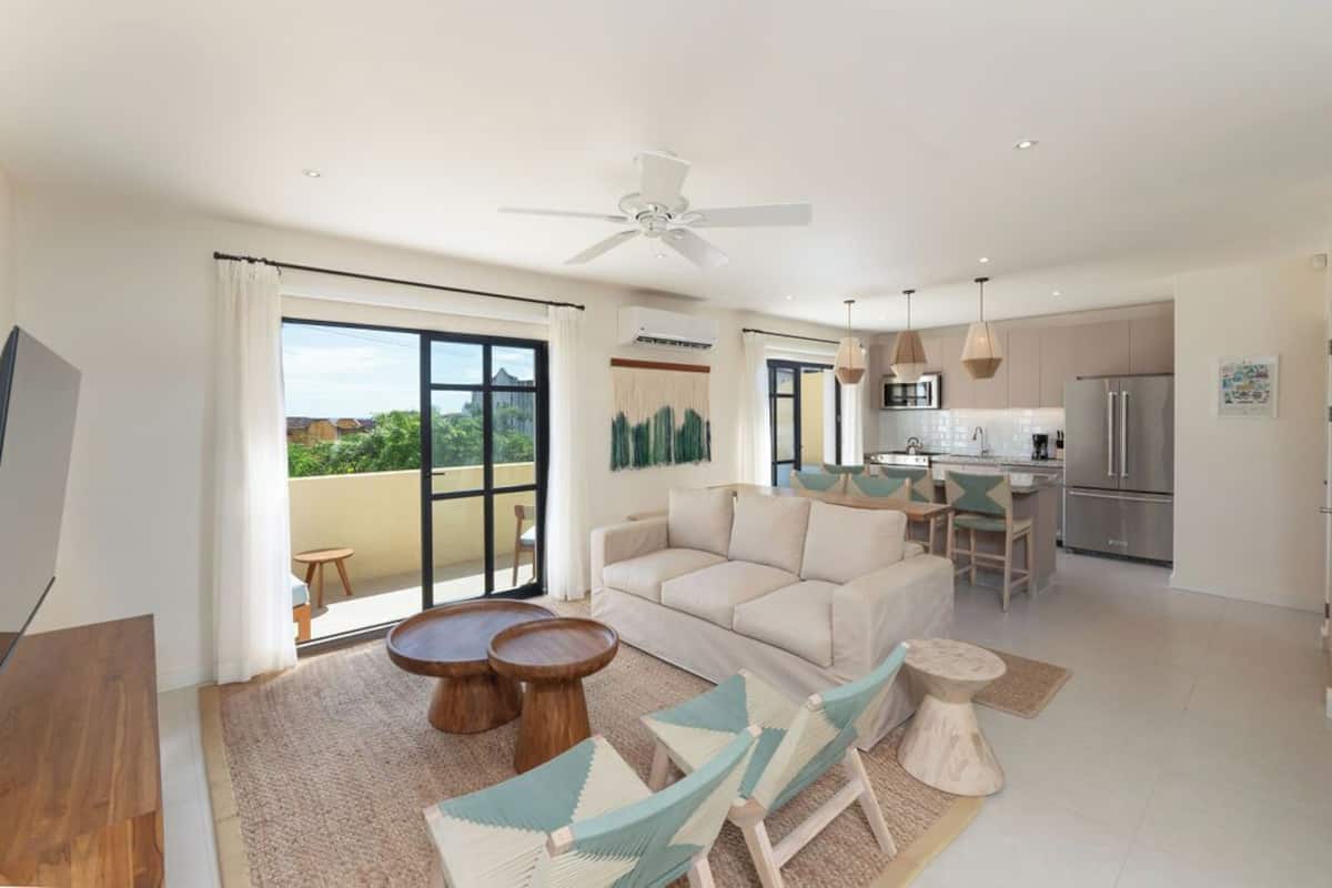Una sala moderna con sofá, mesa de centro y sillas, un balcón con vistas, y una cocina con refrigerador y mesa de comedor.