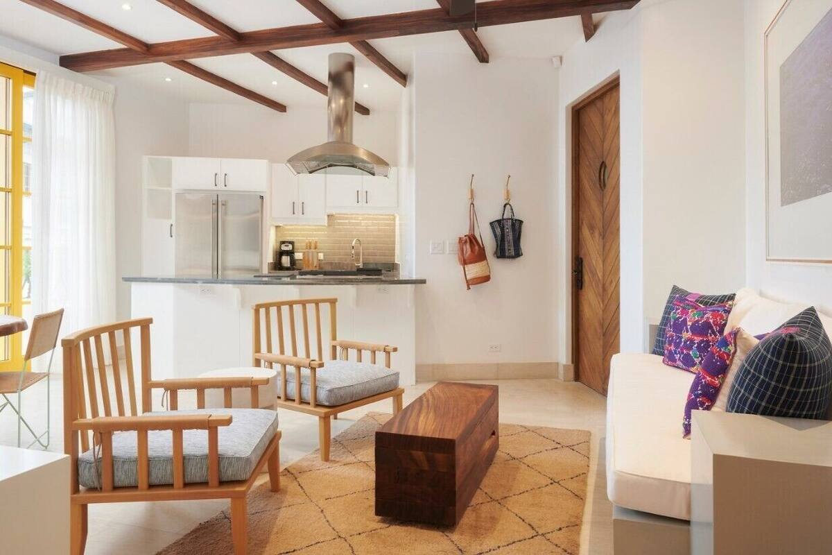 Una sala de estar moderna con un sofá, sillas de madera, una mesita de centro y una cocina al fondo.