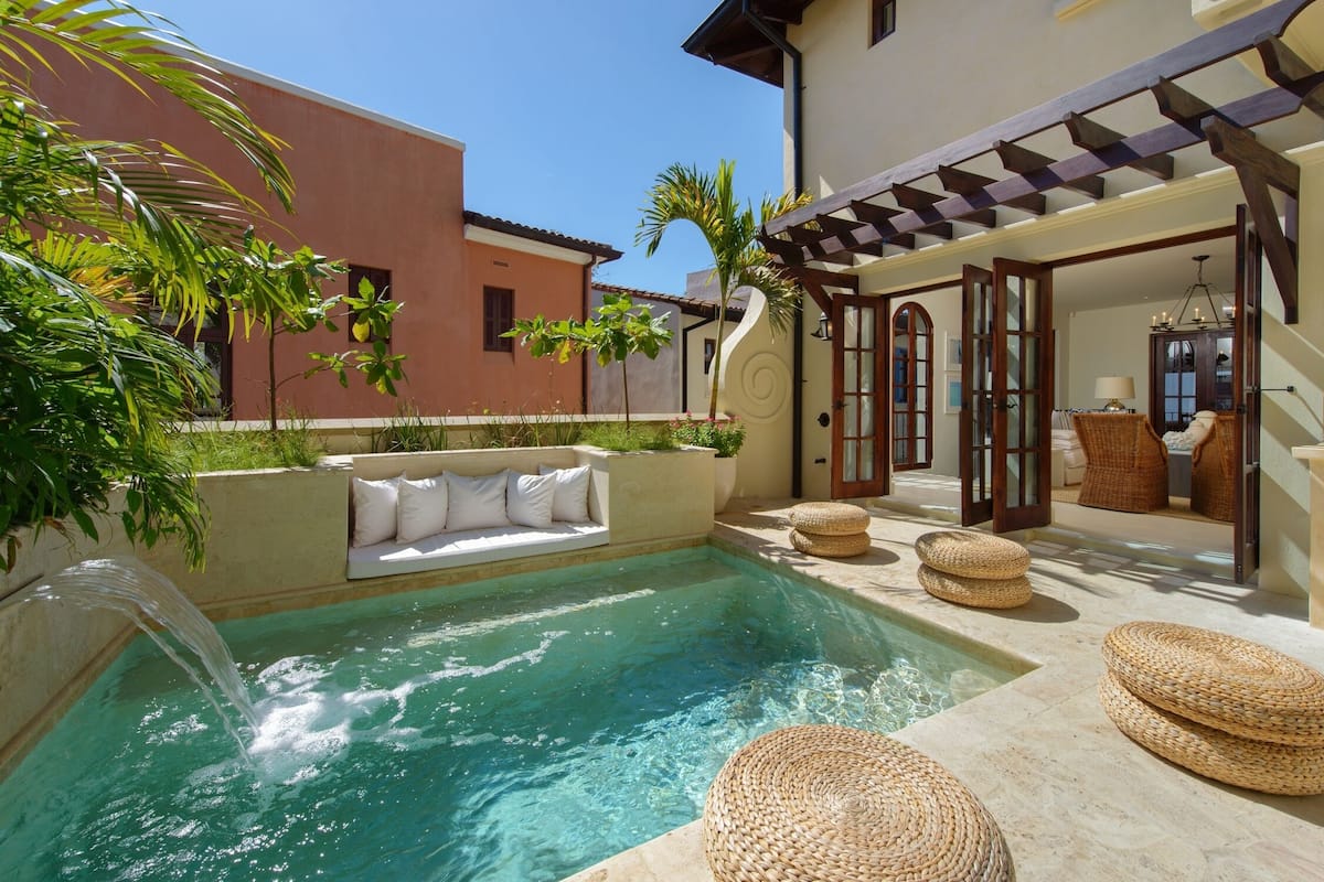 Zona de piscina con un sofá blanco y otomanas tejidas, rodeada por una pérgola y un edificio con una escalera de caracol.
