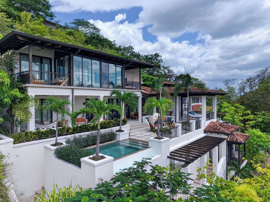 Et moderne hus i to etager med swimmingpool, omgivet af frodig vegetation og en klar himmel.