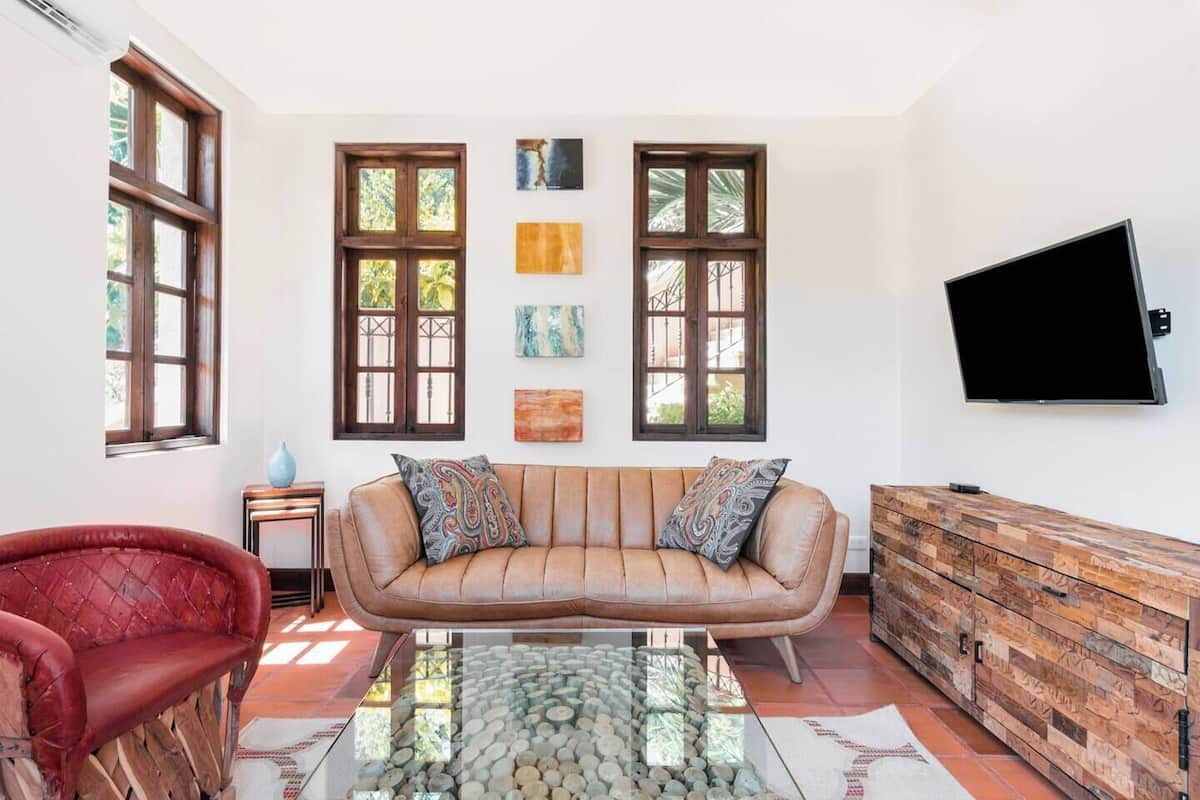 Una sala de estar con un sofá de cuero, un sillón rojo, una mesa de centro de vidrio y un aparador de madera. Hay tres ventanas con marcos de madera y un televisor de pantalla plana montado en la pared.