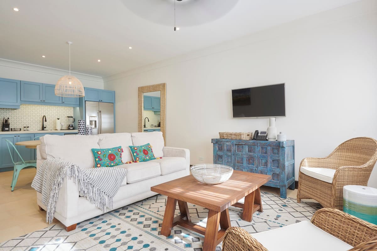 Una sala de estar moderna con un sofá, una mesa de centro y sillas de mimbre. Se ve una cocina con armarios azules y un televisor montado en la pared al fondo.