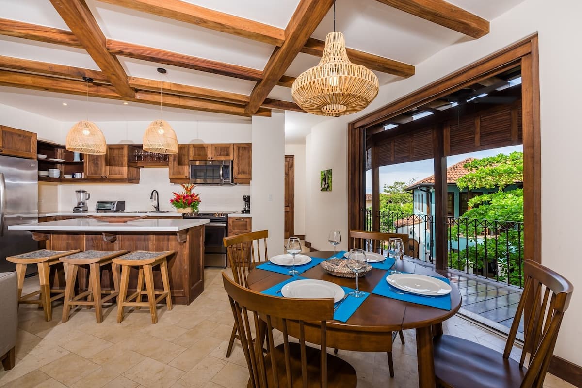 Una cocina moderna con vigas de madera, un comedor con mesa redonda y vistas a un jardín exuberante a través de un ventanal.