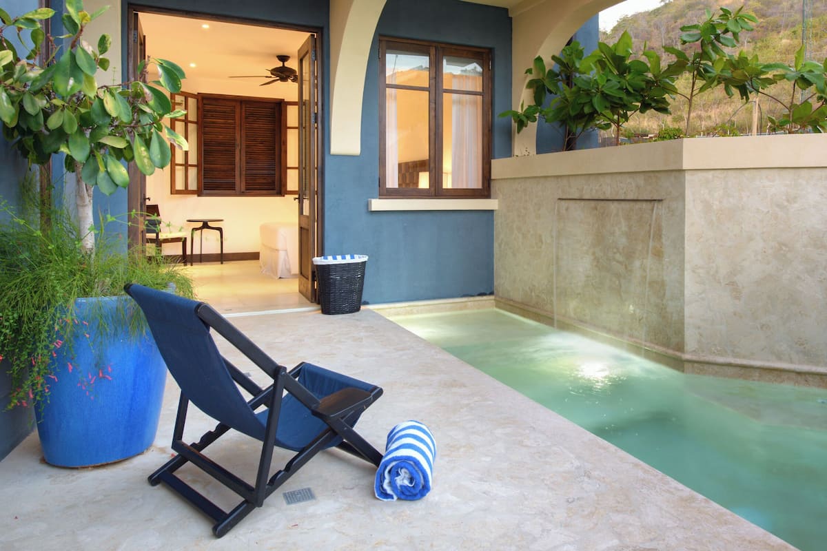 Un área junto a la piscina con una tumbona, una maceta azul y una mesita.