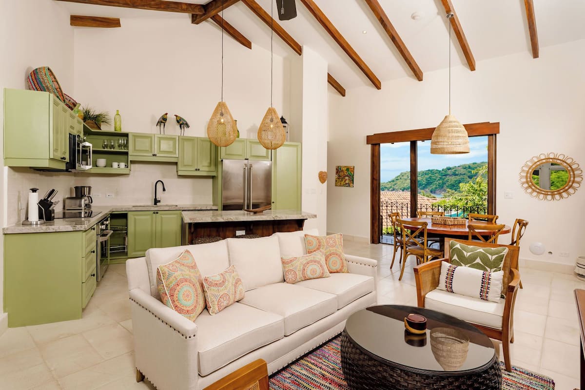 Amplia sala de estar con un sofá blanco, una mesa de comedor con sillas y una cocina con armarios verdes y un ventanal con vistas a un paisaje exuberante.