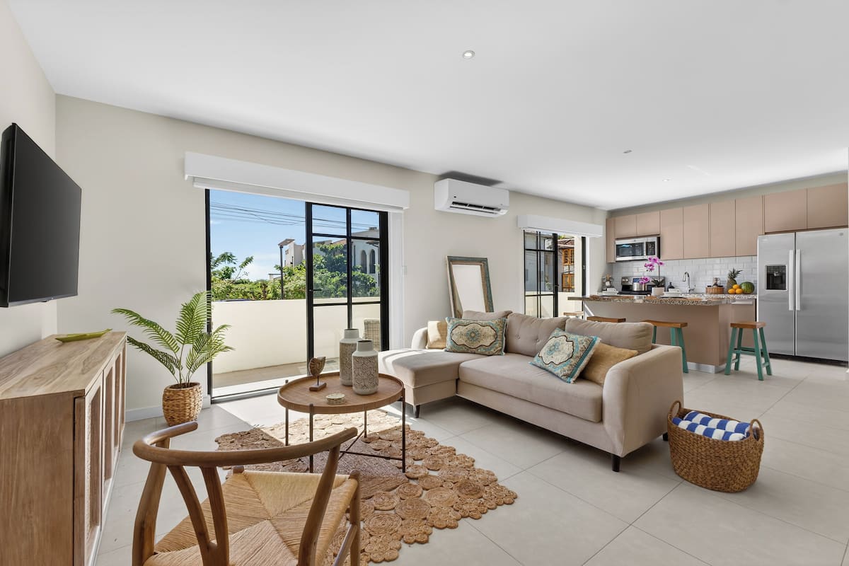 Una sala moderna con un sofá, una mesa de centro y un televisor. Cuenta con una zona de cocina con refrigerador, microondas y una mesa de comedor.