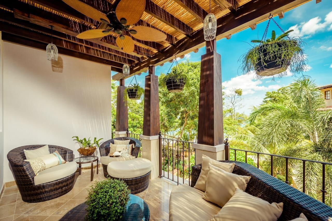 A covered outdoor seating area with wicker furniture, hanging planters, and a view of lush greenery.
