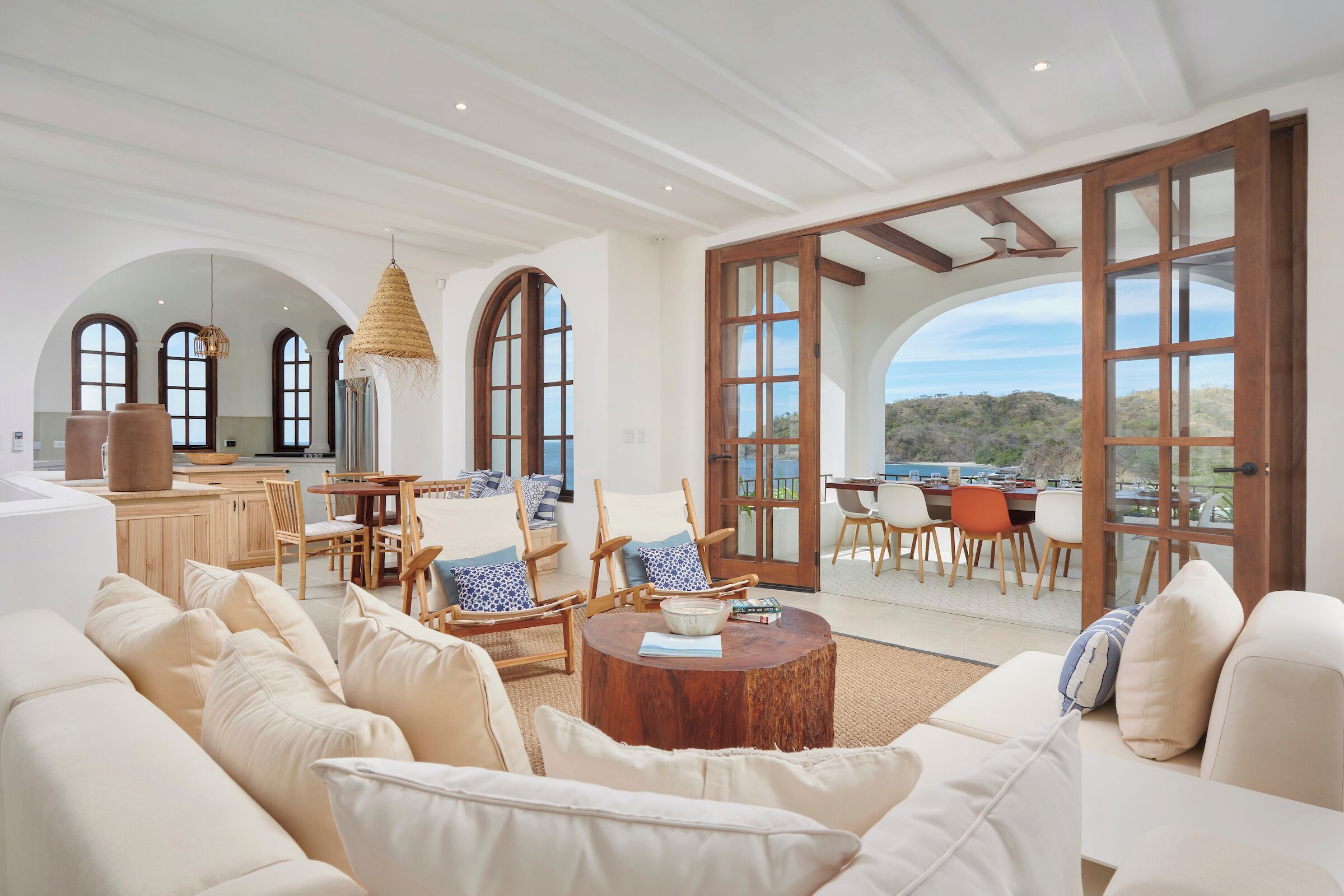 A spacious living area with a large sofa, wooden chairs, a round coffee table, and a view of the outdoors through large windows.