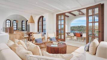 A spacious living area with a large sofa, wooden chairs, a round coffee table, and a view of the outdoors through large windows.