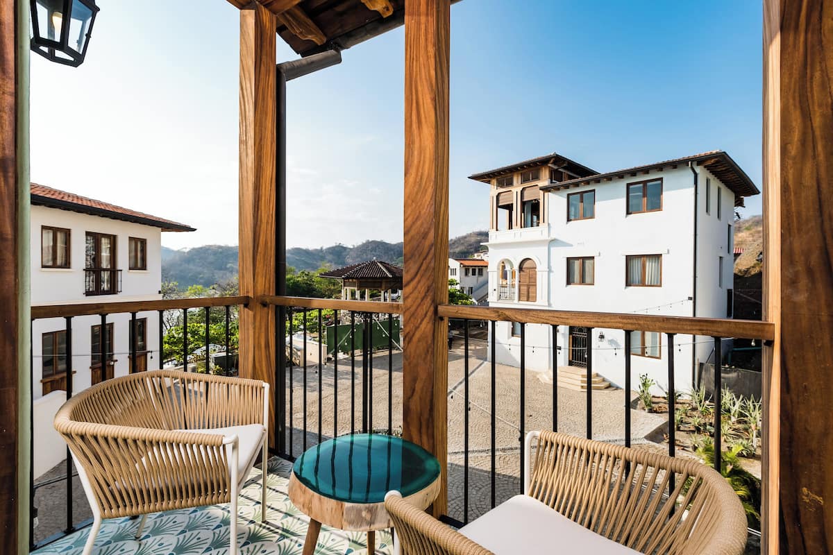 Un balcón con sillas de mimbre y una mesa, con vista a un patio con edificios y una montaña al fondo.