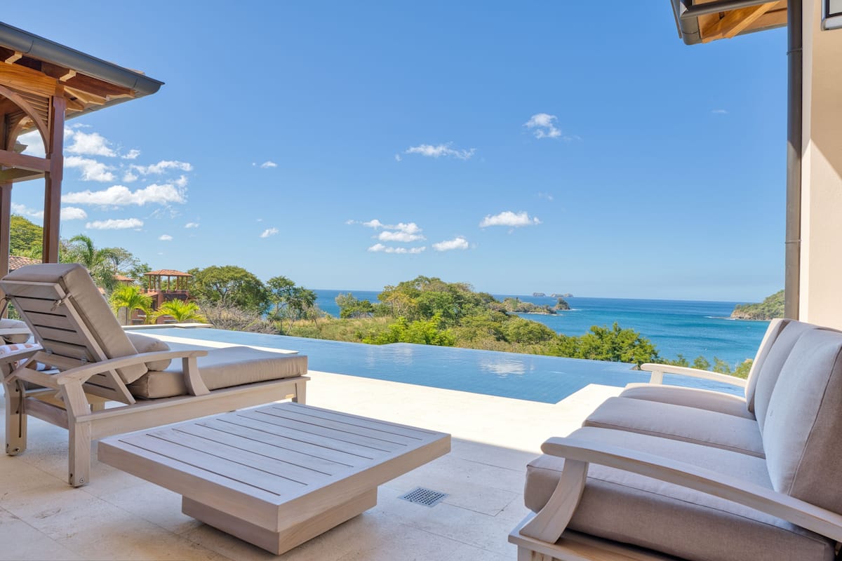 Zona junto a la piscina con sillones de descanso y una mesa, con vistas a un mar azul cristalino y abundante vegetación.