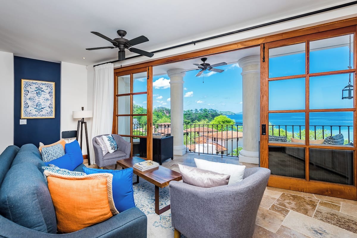 Un salón con sofá, sillón y mesa de centro, ventilador de techo y grandes ventanas con vistas al mar y al verdor.