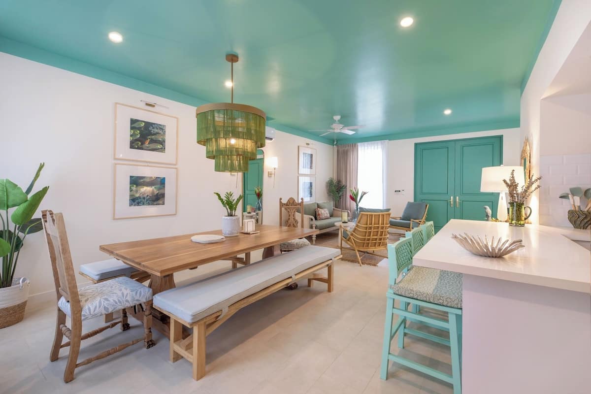 Una sala de estar moderna con una mesa de comedor de madera, un banco y un sofá color turquesa. Hay cuadros en la pared y una lámpara colgante verde grande sobre la mesa.