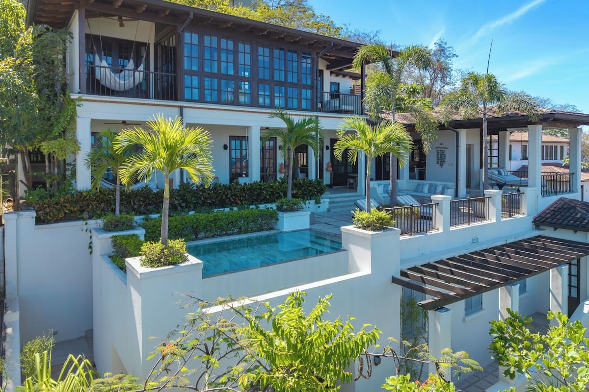 Una casa de dos pisos con piscina, rodeada de abundante vegetación y palmeras.