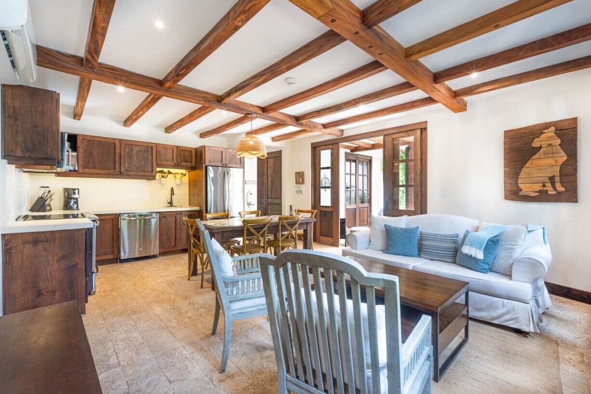 Una cocina amplia con vigas de madera, un comedor con mesa y sillas, y una sala de estar con sofá y un elemento decorativo en la pared.