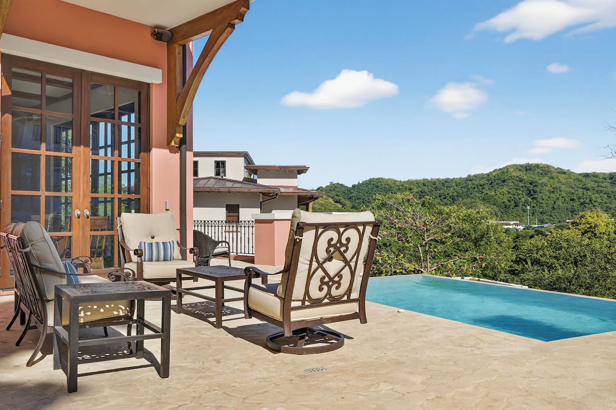 Zona junto a la piscina con mobiliario exterior, vista a la montaña y un edificio con grandes ventanales.