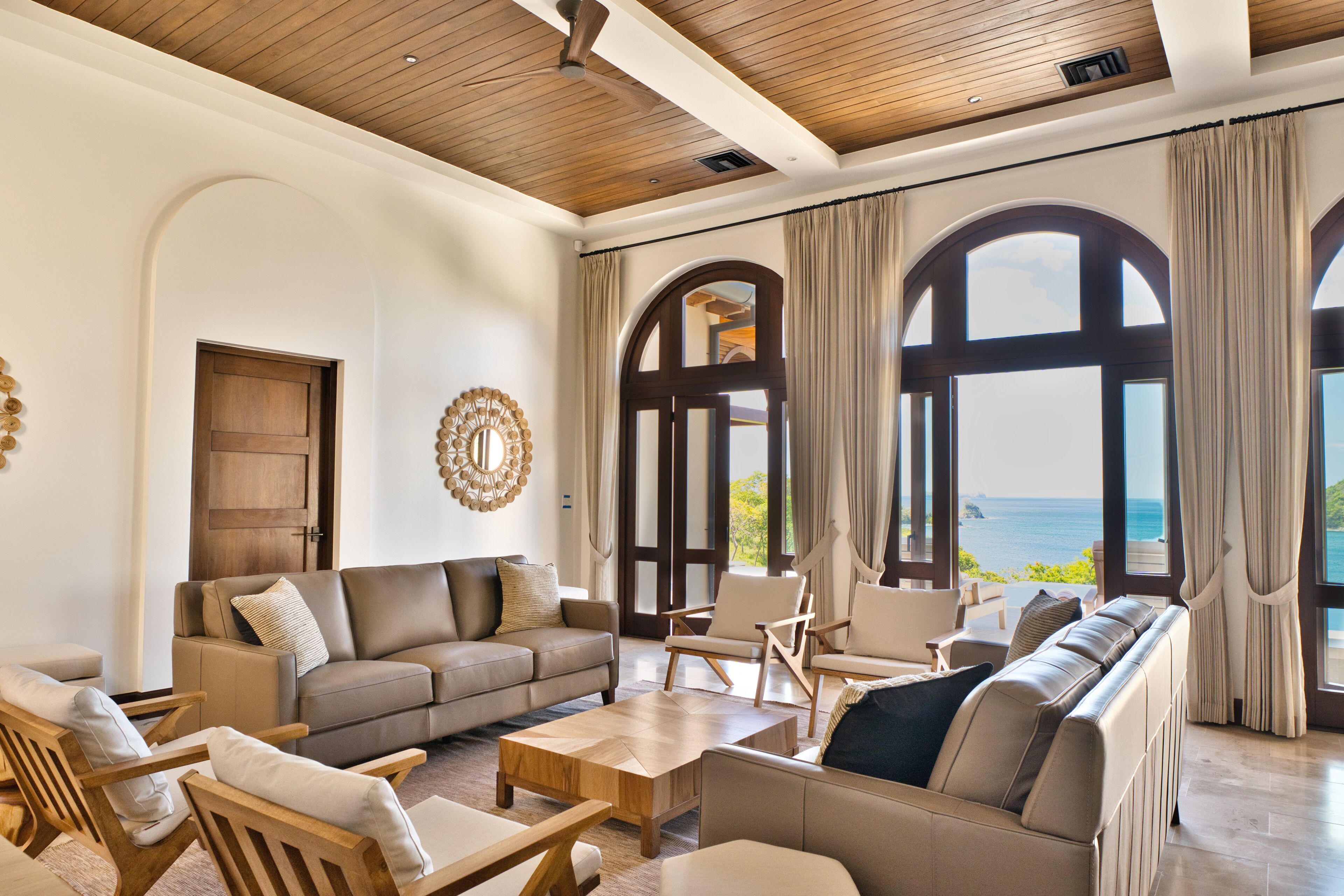 A spacious living area with a large window offering a view of the sea, a wooden ceiling, and a cozy seating arrangement.
