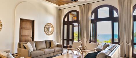 A spacious living area with a large window offering a view of the sea, a wooden ceiling, and a cozy seating arrangement.