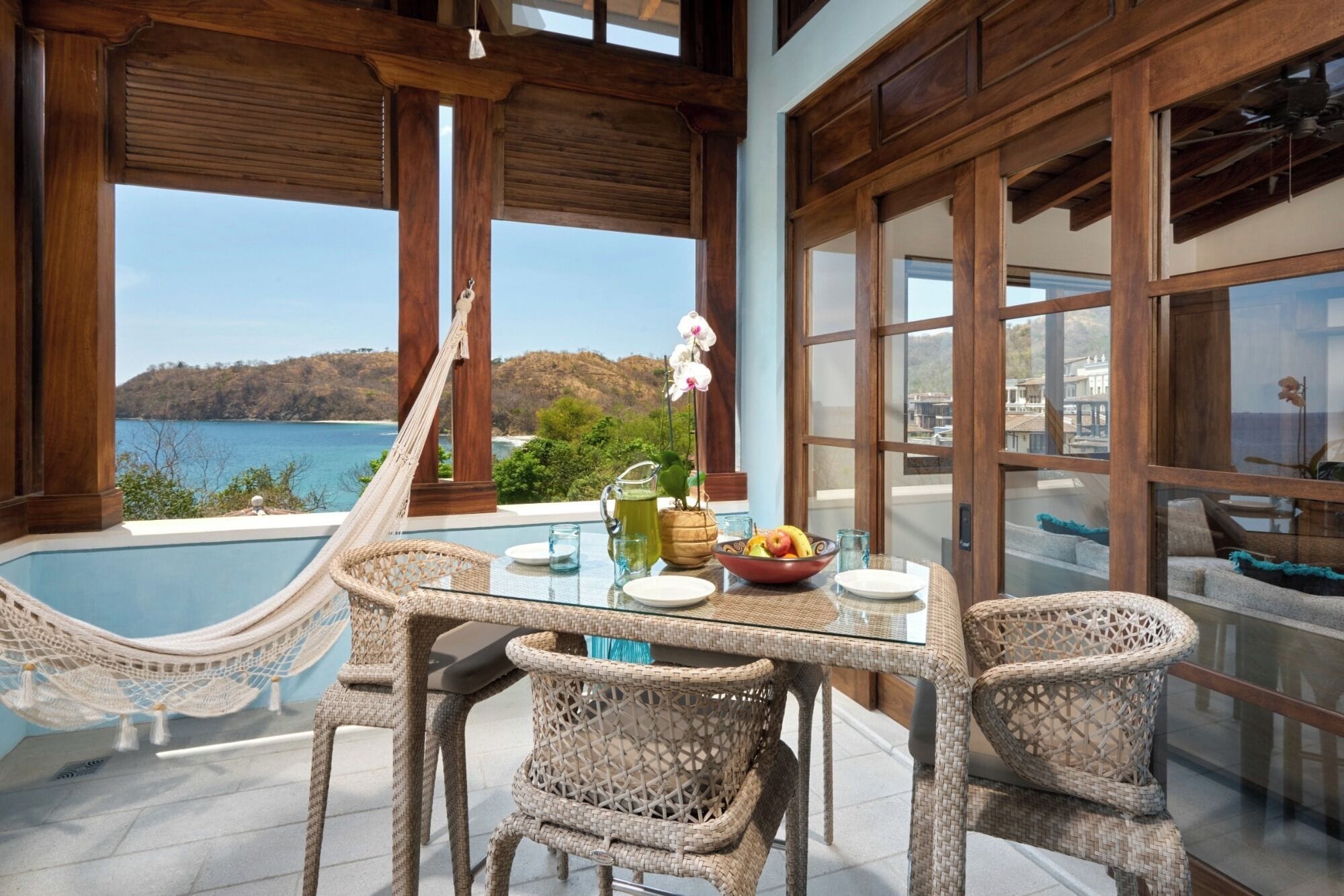 A dining area with a view of the sea, a hammock, wicker chairs, and a table with a pitcher and fruit bowl.