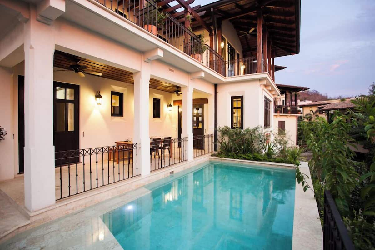 Un área junto a la piscina con una pileta despejada, una zona de estar techada y un balcón con plantas.