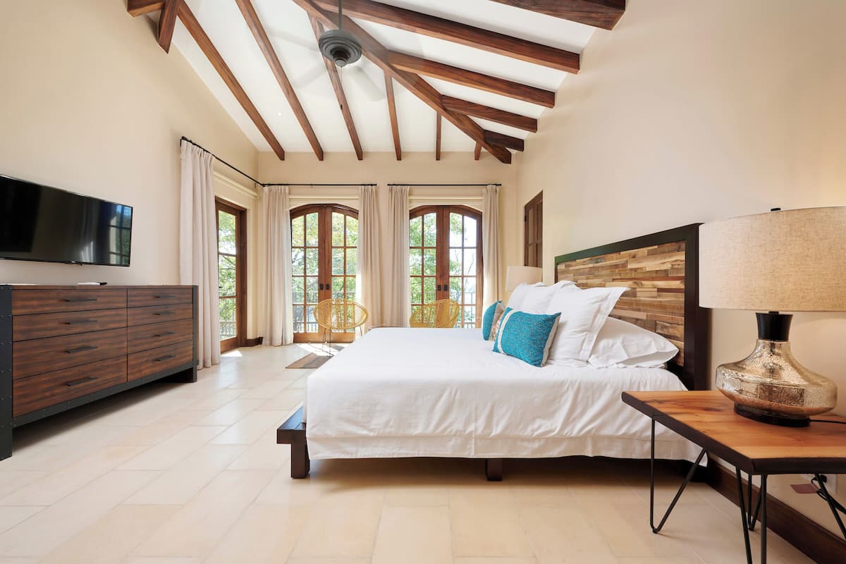 Un dormitorio con una cama grande, cabecera de madera, televisor montado en la pared y una ventana con cortinas.
