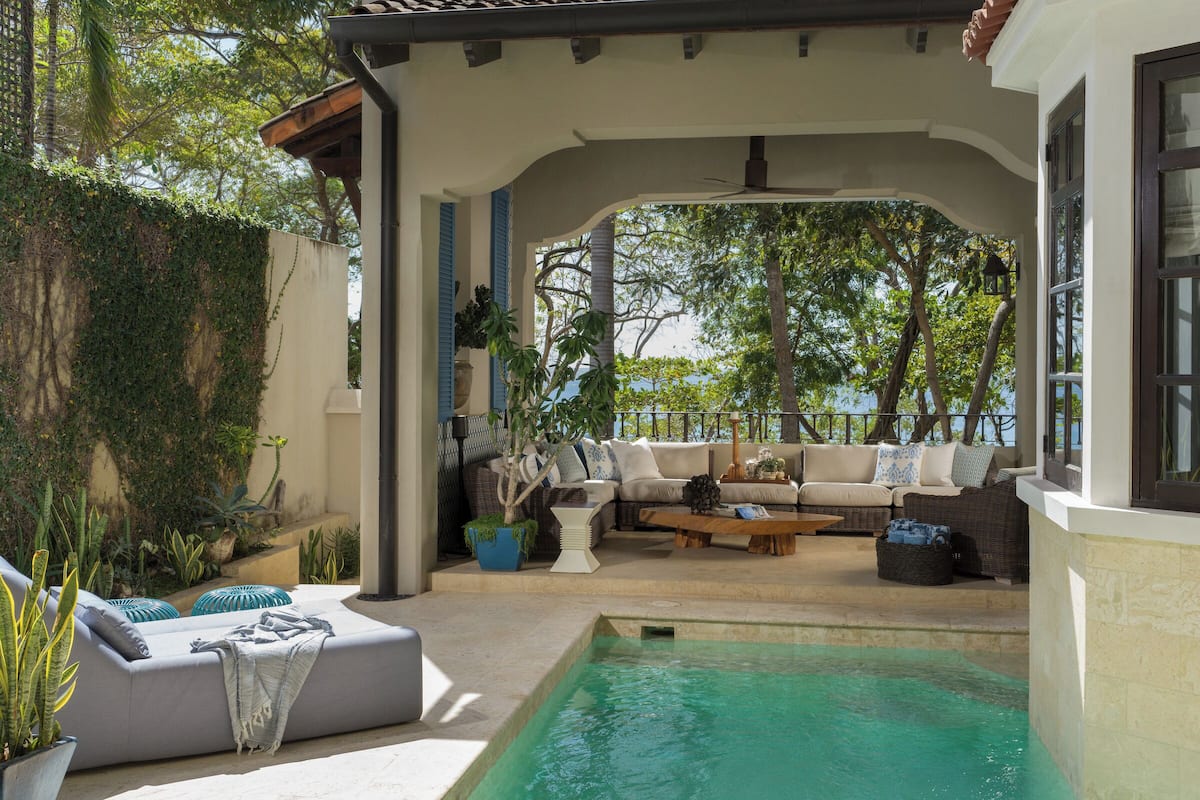 Zona de piscina con espacio de descanso cubierto, piscina y vegetación circundante.