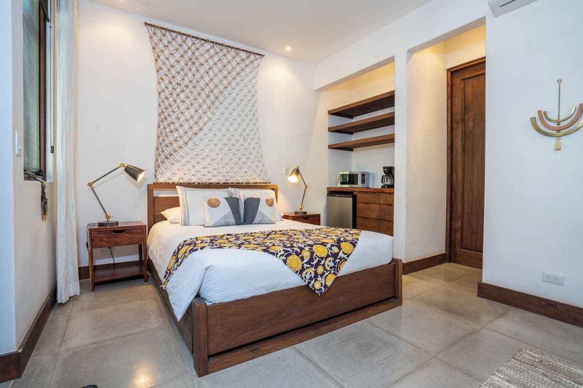 Un dormitorio con cama de madera, mesitas de noche, una mesita auxiliar, estante en la pared, una puerta y un adorno colgante.