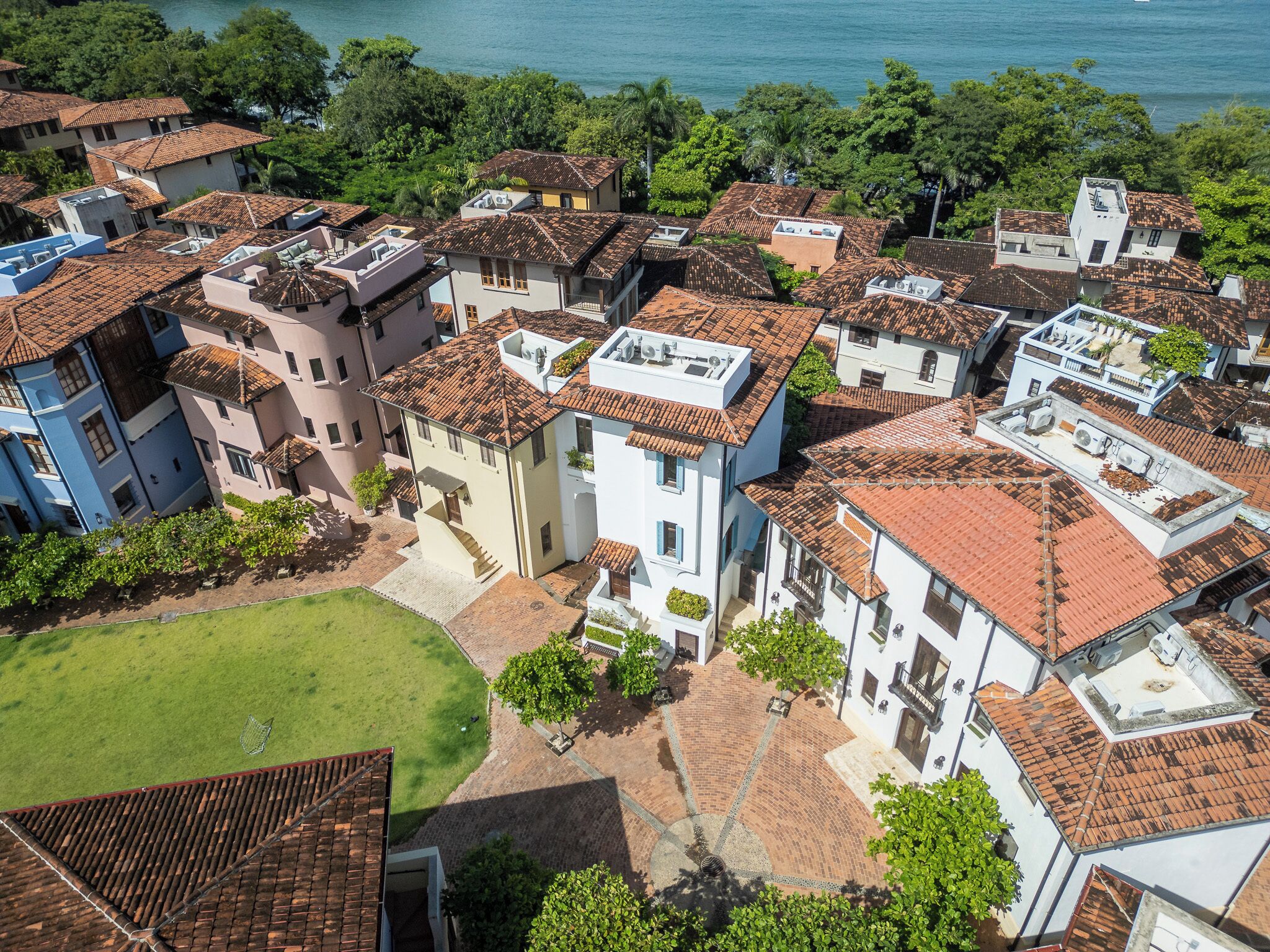 An aerial view of a coastal residential area with terracotta-tiled roofs, white and pastel-colored buildings, and a visible green lawn.